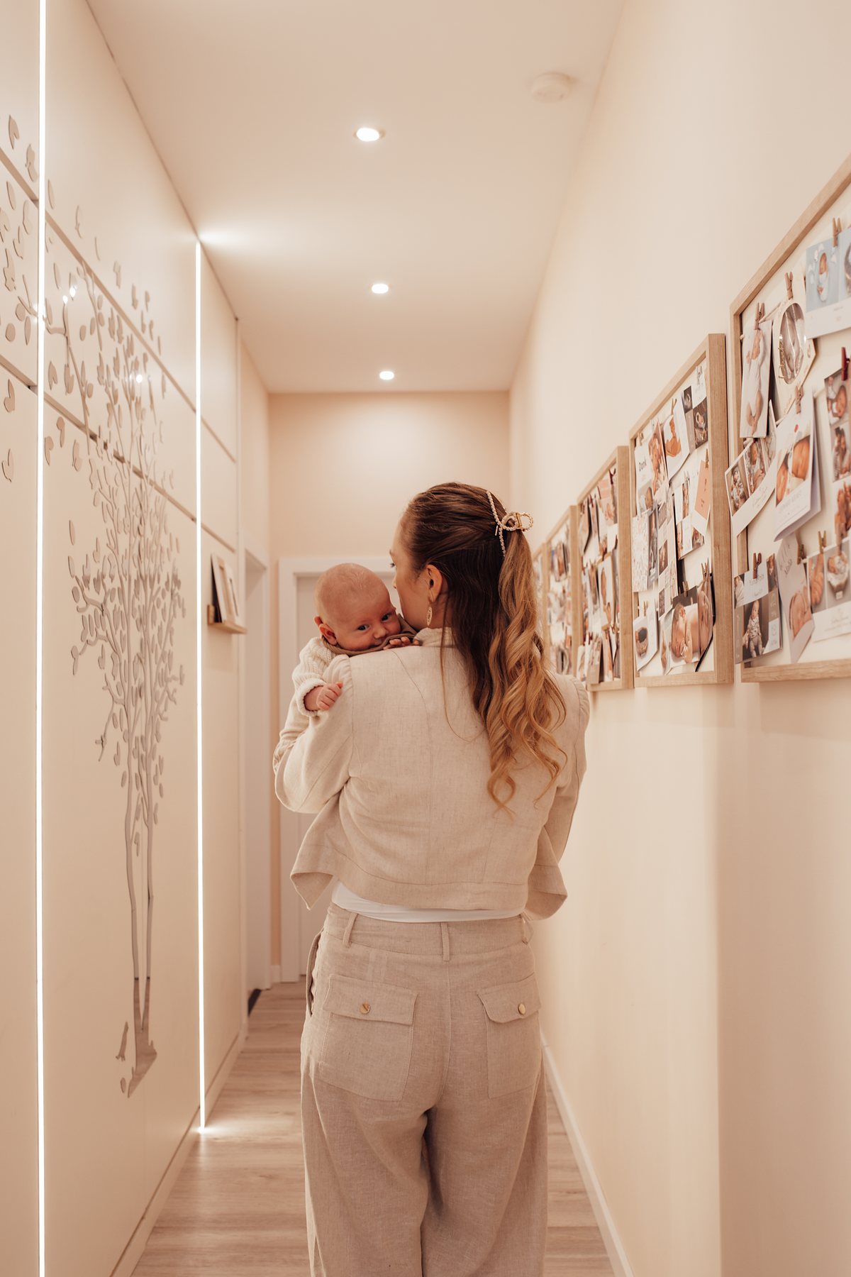 Julia trägt ein Baby durch den beleuchteten Flur der Praxis, vorbei an der Fotowand